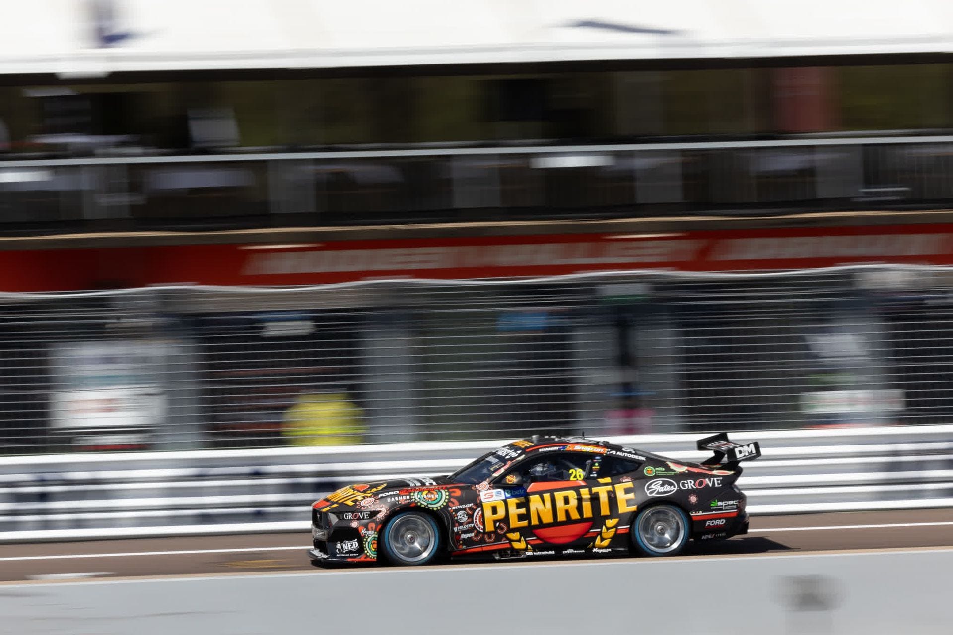 Race car with "Penrite" branding speeds past blurred spectators on a racetrack, showcasing dynamic motion and colorful graphics.