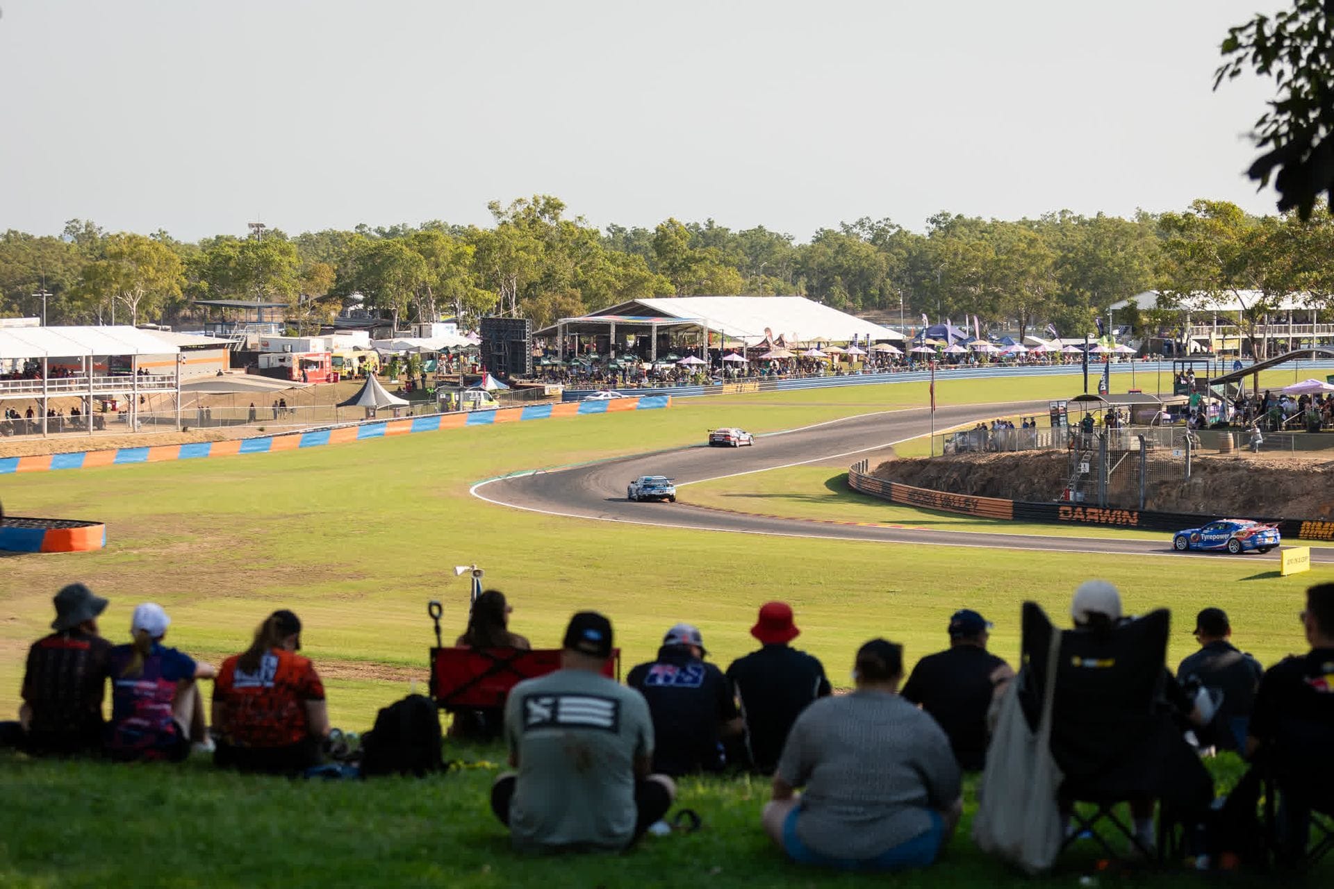 Spectators watch a car race on a sunny day, with cars speeding around a track. Tents and trees are visible in the background.