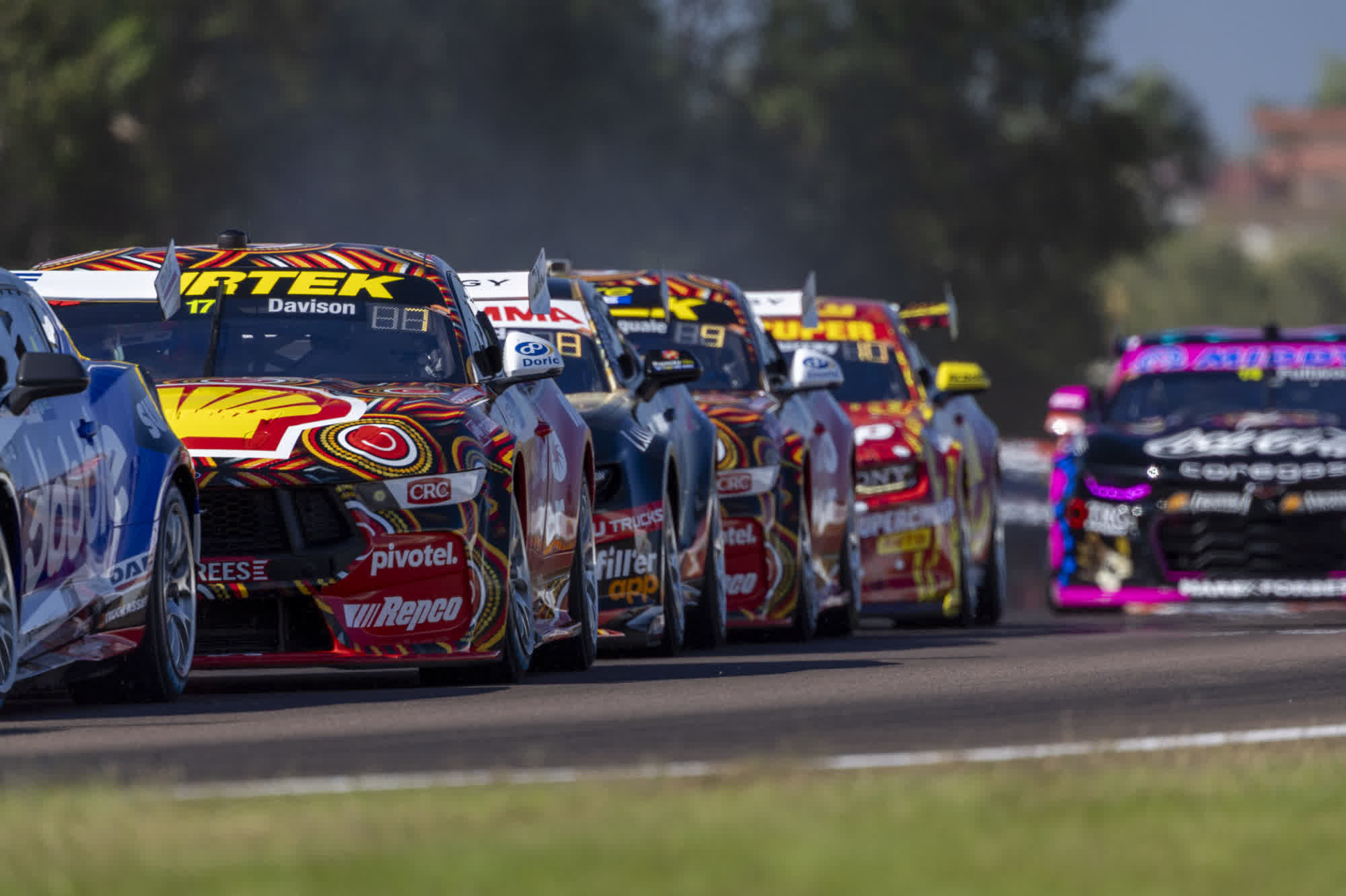Race cars in vibrant colours speed down a track, closely packed, with blurred trees in the background on a sunny day.